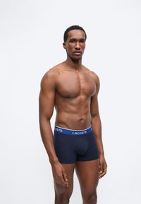 Άνδρας με κανονική φυσική κατάσταση και κοντά μαλλιά στέκεται φορώντας μπλε μποξεράκια Lacoste μπροστά σε ένα απλό φωτεινό φόντο.