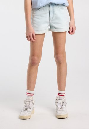 Person wearing light blue rolled-up denim shorts, white high-top sneakers, and white socks with two red stripes, standing against plain background.