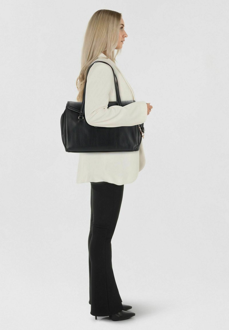 Woman in white blazer and black pants stands sideways holding a large black leather handbag over her shoulder.