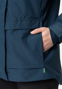 Giacca blu navy con tessuto strutturato, dotata di una grande tasca frontale e un'etichetta verde. Si vede un braccio con una mano parzialmente inserita nella tasca.