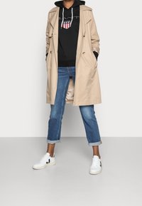 Trench beige sobre una sudadera negra con logo, combinado con unos vaqueros azules arremangados y zapatillas blancas con detalles en negro.