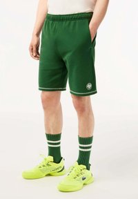 Man i gröna träningsshorts, matchande gröna strumpor med vita ränder och klara gula sportskor från Lacoste.
