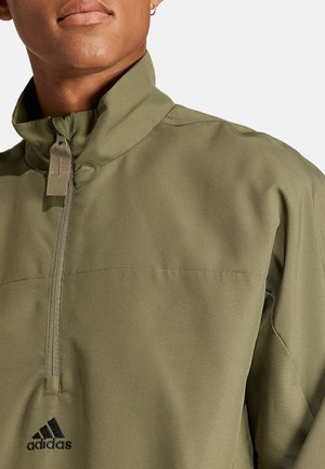 Olijfgroene, lichte half-zip jas met een staande kraag, voorzien van een borstas aan de voorkant en een zwart Adidas-logo nabij de zoom.