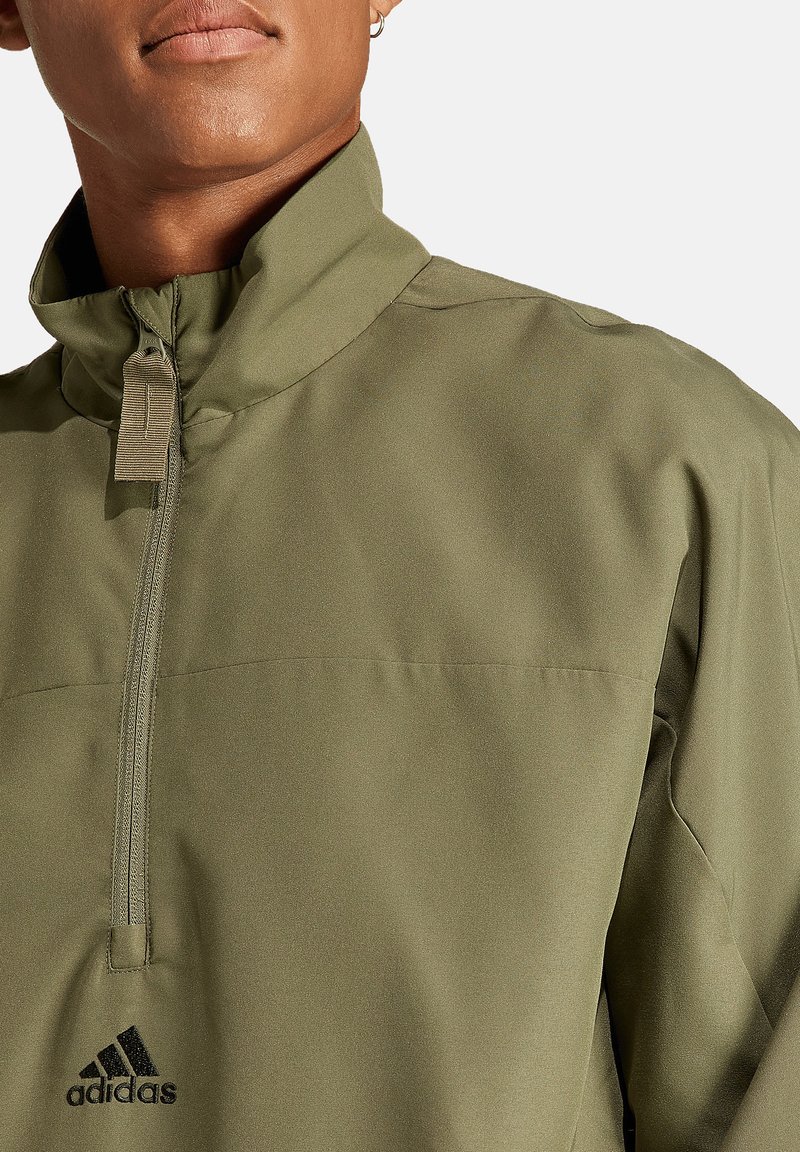 Olivgrüne, leichte Halbzip-Jacke mit Stehkragen, ausgestattet mit einer Brusttasche auf der Vorderseite und einem schwarzen Adidas-Logo nahe dem Saum.