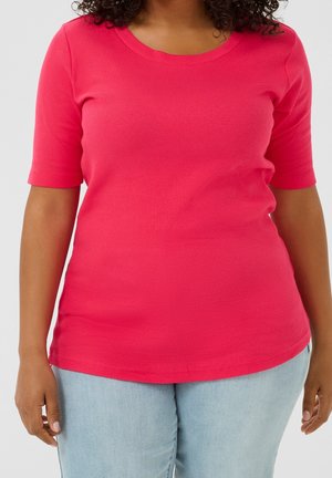 Frau trägt ein knallpinke, kurzärmelige, gerippte Bluse und hellblaue Jeans, gezeigt von den Schultern bis zur Hüfte.