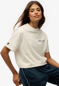 T-shirt en coton crème avec une coupe décontractée, col rond, et "ATHLETICS" imprimé en marine. Présente un logo marine sur la manche gauche.