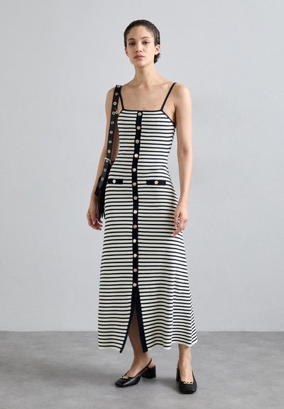 Gestreiftes Strick-Maxikleid mit Spaghetti-Trägern, in Marineblau und Cremefarben, Vorderknöpfen, Seitentaschen und einem Schlitz; kombiniert mit schwarzen Schuhen.