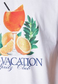 Bílý tričko s výrazným potiskem koktejlové sklenice, pomerančových plátků a zelených listů. Nápis říká "VACATION Spritz Club" modrou barvou.
