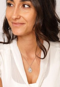 Woman with wavy dark hair wearing a white blouse and a silver pendant necklace with an embossed design.