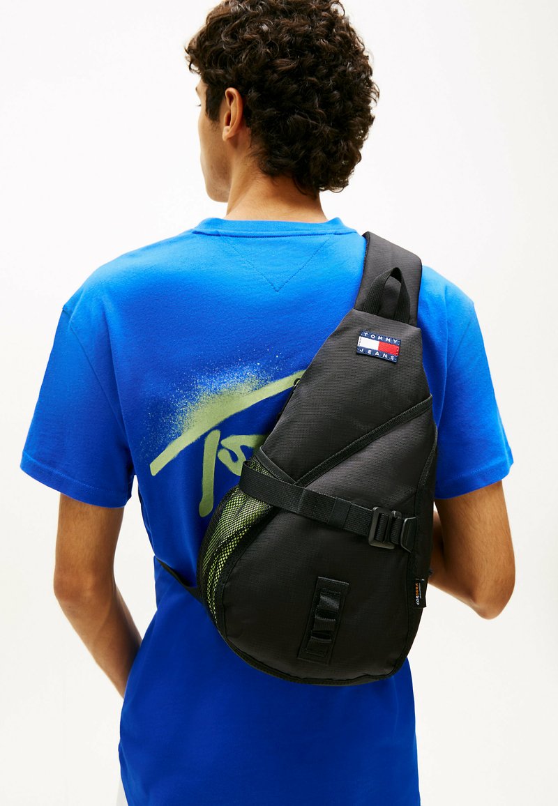 Homme portant un t-shirt bleu à motif graphique avec un sac bandoulière noir Tommy Jeans sur l'épaule, vu de dos sur un fond blanc uni.