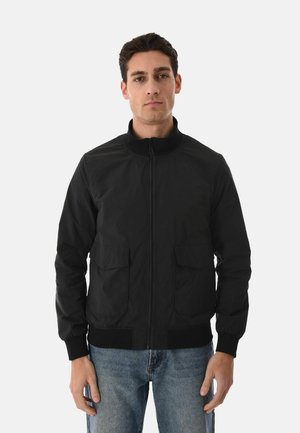 Uomo in piedi di fronte, indossa una giacca nera con zip e tasche frontali e jeans blu, su sfondo neutro.