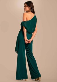 Groene, een-schouder jumpsuit gemaakt van soepel materiaal met wijde benen en zachte draping. Gouden hakken completeren de outfit.