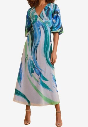 Femme portant une longue robe avec des tourbillons abstraits bleus, verts et blancs et des manches bouffantes, debout dans des talons argentés à bride cheville.