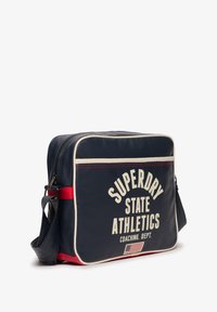 Borsa a tracolla rettangolare blu navy con rifiniture bianche, dettagli rossi e scritta "Superdry State Athletics Coaching Dept." con piccola bandiera americana.