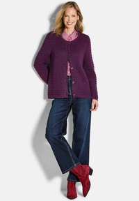 Eine Frau lächelt, trägt einen lila geknöpften Cardigan, ein rosa gemustertes Hemd, dunkle weitgeschnittene Jeans und rote Wildleder-Stiefeletten, während sie mit überkreuzten Beinen steht.