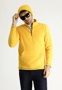 Sudadera de forro polar amarillo con cuello de media cremallera, bolsillo delantero y puños de canalé, combinada con un gorro amarillo a juego y gafas de sol negras.