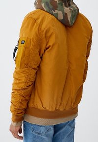 Veste bomber jaune avec ourlet et poignets côtelés, avec une capuche camouflage. Détails comprenant une poche zippée sur la manche gauche.