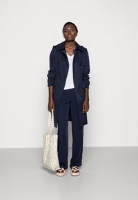 Navy trench coat with double-breasted buttons, paired with a white v-neck shirt and navy trousers. Accented by a patterned beige tote and shoes.