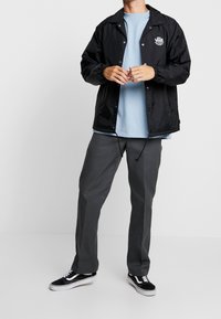 Giacca a vento nera in nylon con chiusura a scatto, dotata di polsini elastici e logo Vans. Indossata sopra una maglietta blu chiaro e pantaloni grigi.