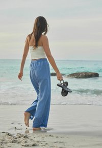 Pantaloni ușor albastri, lejeri, cu talie elastică, purtați cu un top alb, ajustat. Ținând sandale negre flip-flop, mergând pe nisip umed.