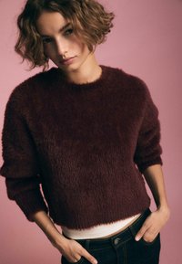 Burgunderfarbener, flauschiger Pullover mit Rundhalsausschnitt und drei Viertellängenärmeln, über einem weißen Tanktop getragen und kombiniert mit dunklen Jeans.