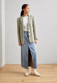 Blazer à carreaux verts, haut ajusté blanc, jupe en jean taille haute avec fente, bottes beiges à bout en caoutchouc, fond en parquet.