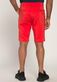 Rode shorts van een gladde, lichte stof met een achterzak aan elke kant. Draagt met zwarte schoenen, weergegeven vanaf de achterkant.
