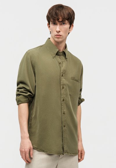 Νέος άνδρας με κοντά καστανά μαλλιά φοράει πουκάμισο μακρυμάνικο σε χακί πράσινο με κουμπιά και μανίκια γυρισμένα, καθώς και ανοιχτό μπεζ παντελόνι, πάνω σε λευκό φόντο.