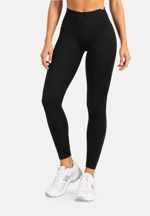 Mujer con leggings negros de talle alto y zapatillas deportivas blancas, de pie con las manos relajadas a los lados sobre un fondo liso.