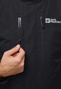 Veste imperméable noire avec deux poches poitrine zippées, texture lisse, et logo Jack Wolfskin sur le côté gauche.