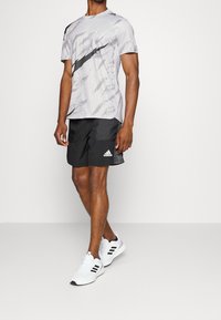 Camiseta atlética gris con un patrón y una franja diagonal negra, combinada con pantalones cortos negros con bolsillos laterales y zapatillas deportivas blancas.