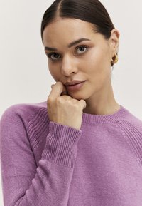 Lila gestrickter Pullover mit gerippten Bündchen und Rundhalsausschnitt, vor einem neutralen Hintergrund, mit einer glatten Textur und lockerer Passform.