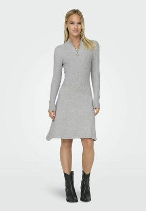 Robe tricotée grise avec des manches longues côtelées et un col en V, se transformant en une bordure évasée. Associée à des bottines noires.