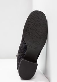Bottine noire à semelle texturée, talon bloc, fermeture éclair latérale et tissu avec motif floral en relief, vue par le dessous.