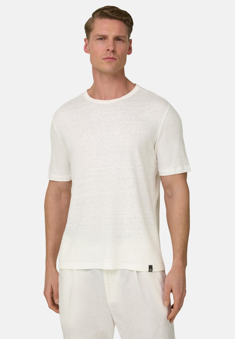 Man draagt een effen witte T-shirt met korte mouwen en ronde hals en witte broek, staand tegen een effen lichte achtergrond.