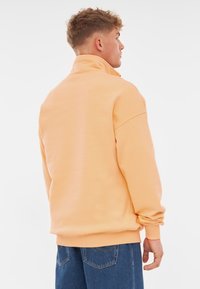 Lichtoranje pullover met hoge kraag, oversized pasvorm, geribde manchetten en tailleband. Combinatie met losse blauwe spijkerbroek.