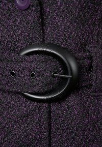 Tejido texturizado en púrpura y negro con un detalle de hebilla negra circular y una correa con aspecto de cuero que presenta pequeños agujeros para ajustes.