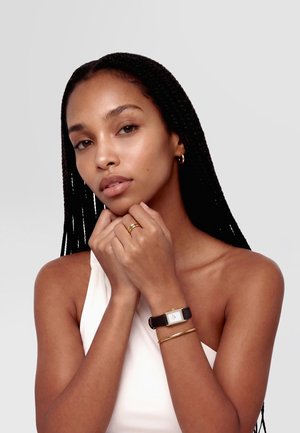 Femeie cu cozi împletite lungi, purtând un top alb, cercei mari de aur, inele de aur, o brățară de aur și un ceas dreptunghiular cu curea neagră din piele.