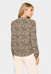 Blouse à imprimé léopard avec des manches longues, en tissu léger. Présente un ourlet arrondi et des poignets élastiques, dans des couleurs beige, marron et noir.