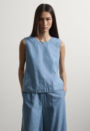 Vrouw met lang donker haar, gekleed in een mouwloze lichtblauwe linnen top en bijpassende wijde broek, staand met handen in de zakken.