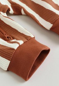 Suurelt plaanilt pruunide ja valgete soonikkoeliste varrukate lähedalt, millel on pruunid elastsed mansetid ja ühe varruka peal väike tõmblukk.