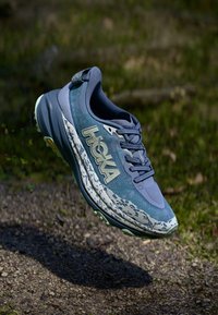 Chaussure de course HOKA en bleu et vert avec une tige texturée, une semelle intermédiaire blanche ornée d'un motif éclaboussé, et une semelle extérieure améliorant l'adhérence.