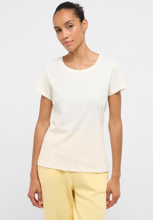 Woman with dark hair in a bun wearing a plain white short-sleeve T-shirt and light yellow pants, standing against a light background.