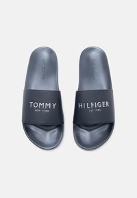 Navy blue slide sandals made of smooth synthetic material. Features branded text in silver: "TOMMY NEW YORK" and "HILFIGER EST. 1985."