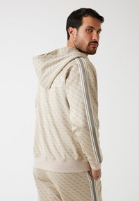 Sudadera beige con un patrón integral y acentos a rayas blancas en las mangas. Cuenta con una capucha con cordones y puños y dobladillo acanalados.