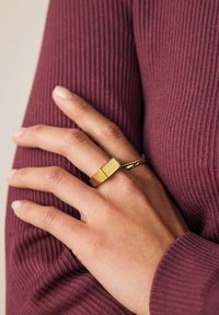 Goldring mit rechteckigem Design, das eine glatte Oberfläche und ein strukturiertes Drehdetail aufweist, getragen an einer Hand, die sich gegen einen gerippten weinrotfarbenen Stoff lehnt.