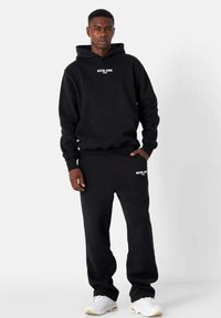 Sudadera negra con capucha y pantalones de chándal a juego, hechos de una tela suave, con un logo en el pecho y en el muslo. Diseño sencillo con puños de canalé.