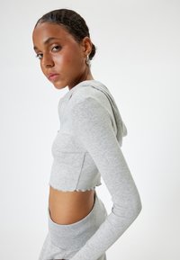 Grijze cropped hoodie van ribbelstof, met lange mouwen, een afgeronde zoom en een capuchon. Opvallende uitsparingen aan de zijkanten verbeteren het ontwerp.