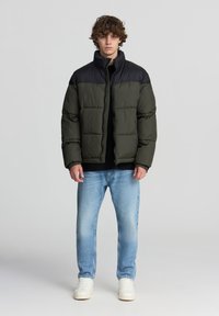 Giubbotto imbottito con sezione superiore nera e parte inferiore verde scuro, dotato di chiusura con zip, forma ampia e texture trapuntata. Indossato con jeans chiari.