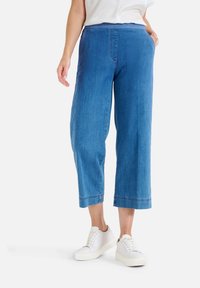 Pantalons cropped en denim bleu clair avec une coupe décontractée, détails de poches et ceinture, associés à des baskets blanches sur un fond uni.